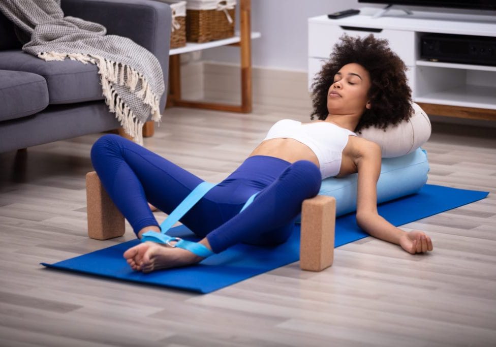 Relaxed Young Woman Lying On Fitness Mat Doing Exercise With Yoga Belt And Two Blocks